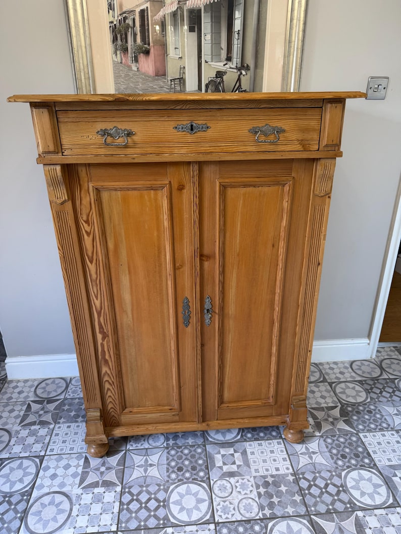 May include: A light brown wooden cabinet with two doors and a drawer. The cabinet has ornate metal handles and a keyhole. The wood grain is visible. The cabinet is on a patterned tile floor.