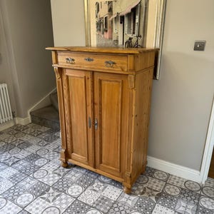 May include: A light brown wooden cabinet with two doors and a drawer, featuring ornate metal hardware. The cabinet is set against a neutral wall, with a framed picture above it. The cabinet has a classic design and is likely used for storage.