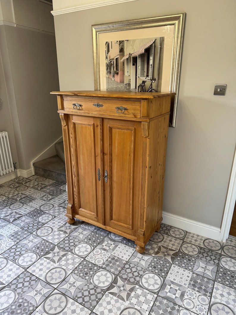 May include: A light brown wooden cabinet with two doors and a drawer, featuring ornate metal hardware. The cabinet is set against a neutral wall, with a framed picture above it. The cabinet has a classic design and is likely used for storage.