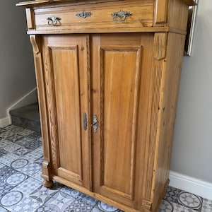 May include: A light brown wooden cabinet with two doors and a drawer. The cabinet features decorative metal handles and drawer pulls. The wood grain is visible, and the cabinet rests on short, rounded feet. The cabinet is likely used for storage.