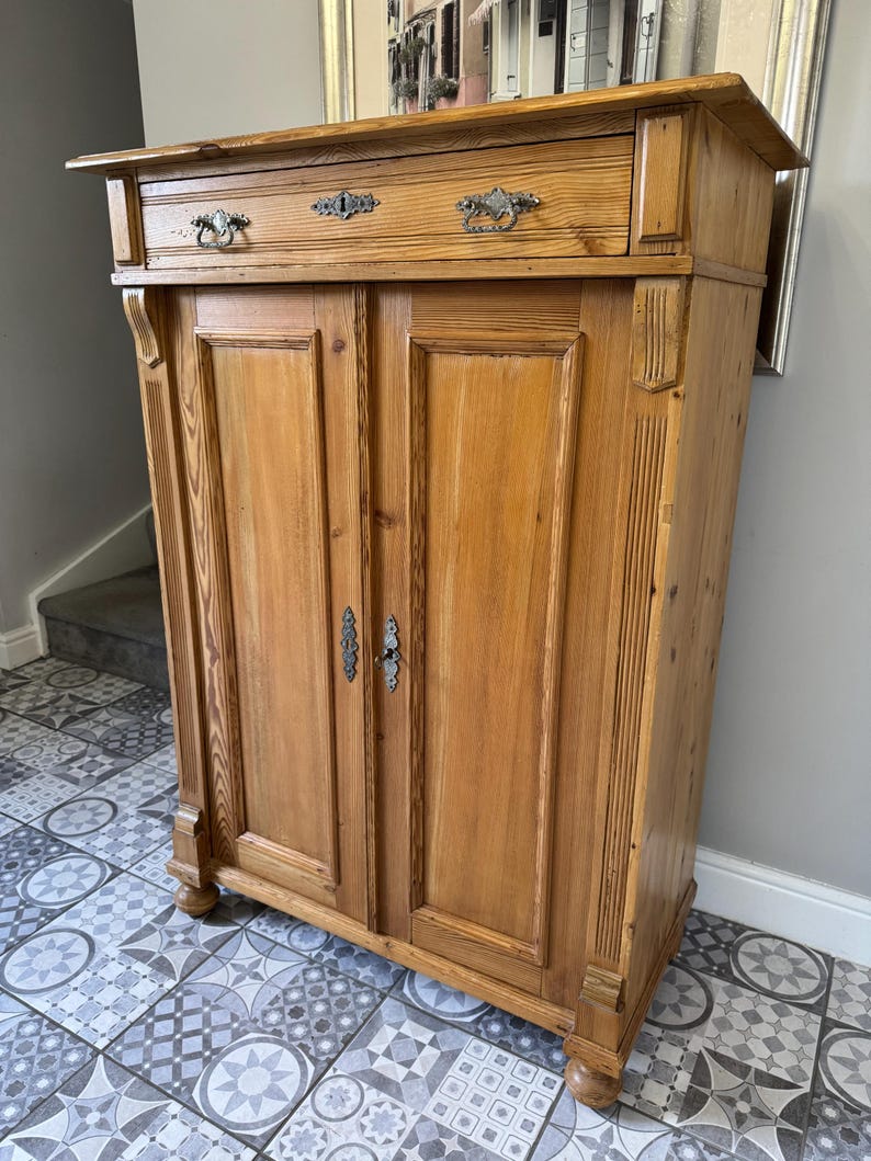 May include: A light brown wooden cabinet with two doors and a drawer. The cabinet features decorative metal handles and drawer pulls. The wood grain is visible, and the cabinet rests on short, rounded feet. The cabinet is likely used for storage.