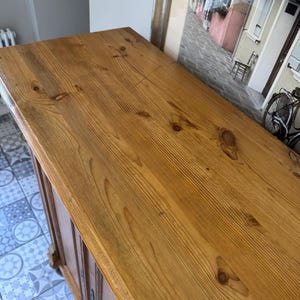 May include: A light brown wooden cabinet with a varnished finish. The top surface is made of planks with visible wood grain and knots. The cabinet has a classic design and appears to be vintage or antique.