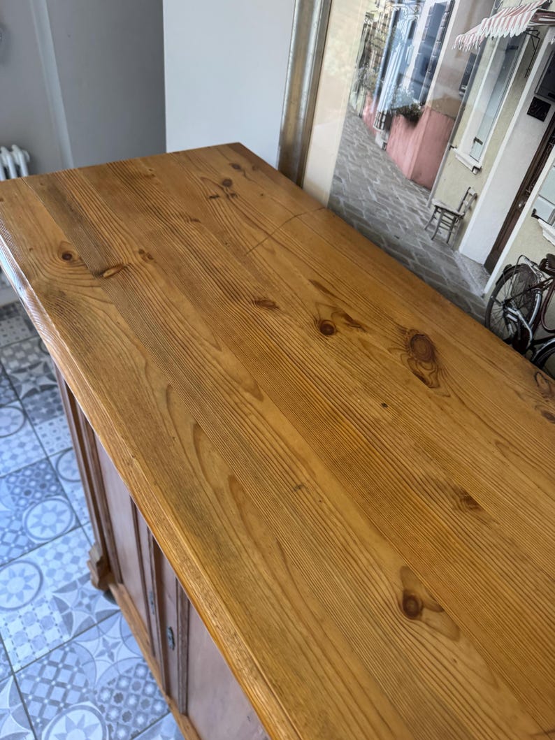 May include: A light brown wooden cabinet with a varnished finish. The top surface is made of planks with visible wood grain and knots. The cabinet has a classic design and appears to be vintage or antique.
