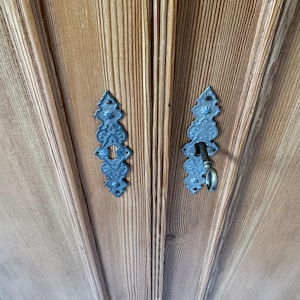 May include: Close-up of a wooden cabinet door with two ornate, dark metal keyhole covers. One cover has a key inserted. The wood grain is visible, and the cabinet has a light brown colour.
