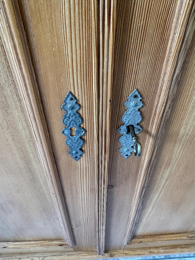 May include: Close-up of a wooden cabinet door with two ornate, dark metal keyhole covers. One cover has a key inserted. The wood grain is visible, and the cabinet has a light brown colour.