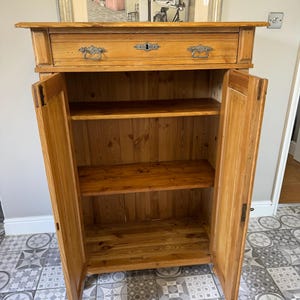 May include: A light-coloured wooden cabinet with two doors open, revealing shelves inside. The cabinet has a drawer at the top with ornate metal handles and a keyhole. The wood grain is visible, and the cabinet appears to be vintage or antique.