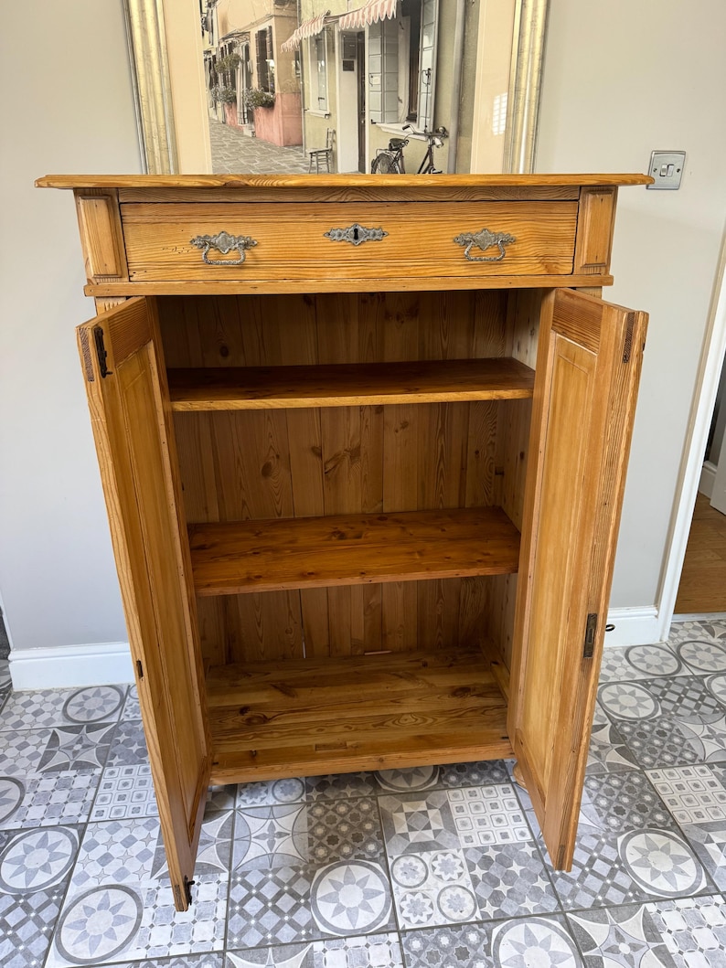 May include: A light-coloured wooden cabinet with two doors open, revealing shelves inside. The cabinet has a drawer at the top with ornate metal handles and a keyhole. The wood grain is visible, and the cabinet appears to be vintage or antique.