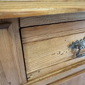 May include: Close-up of a wooden cabinet with a drawer. The cabinet is made of light-coloured wood with visible grain. The drawer has a decorative metal handle. The cabinet appears to be vintage or antique.