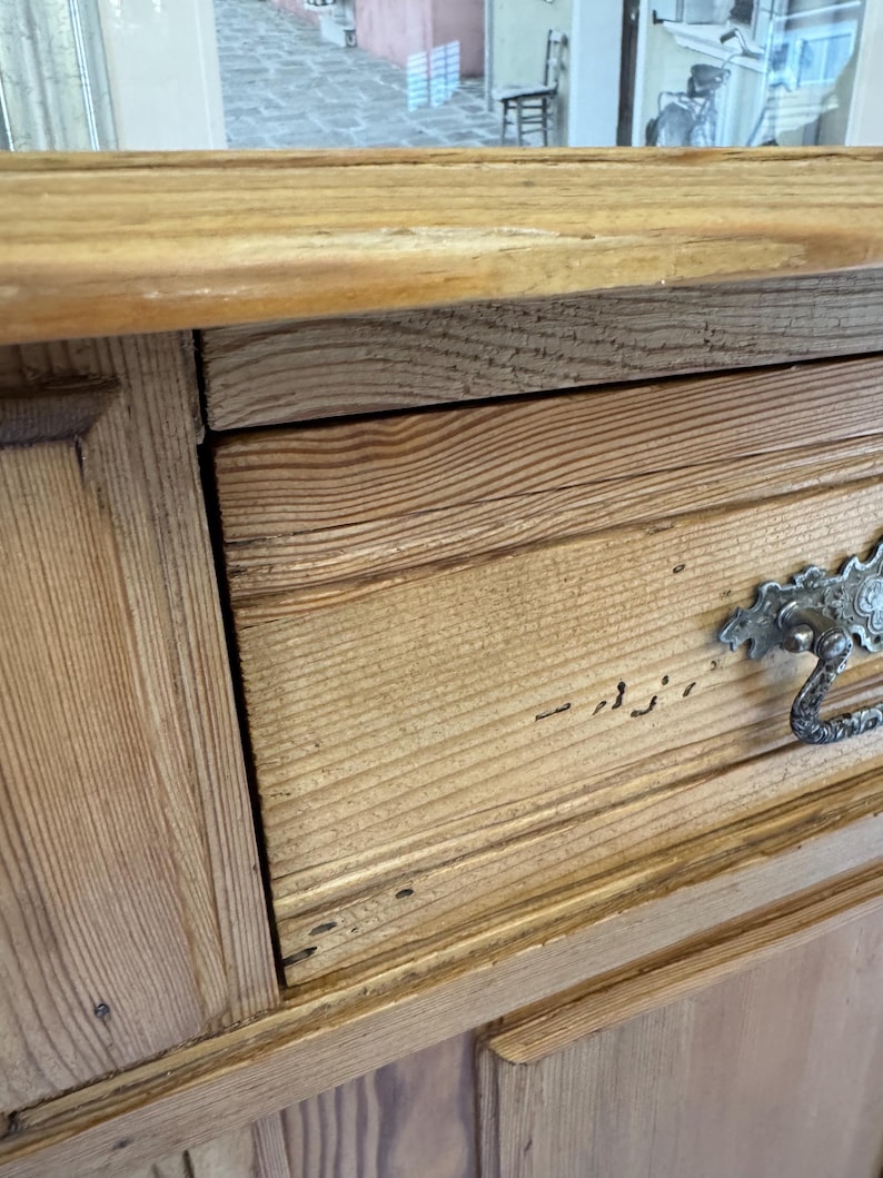 May include: Close-up of a wooden cabinet with a drawer. The cabinet is made of light-coloured wood with visible grain. The drawer has a decorative metal handle. The cabinet appears to be vintage or antique.