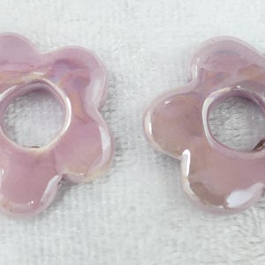 May include: Two iridescent, light purple flower-shaped ceramic beads. Each bead has a central hole and five rounded petals. The beads have a glossy finish and reflect light, creating a shimmering effect. They are set against a white background.