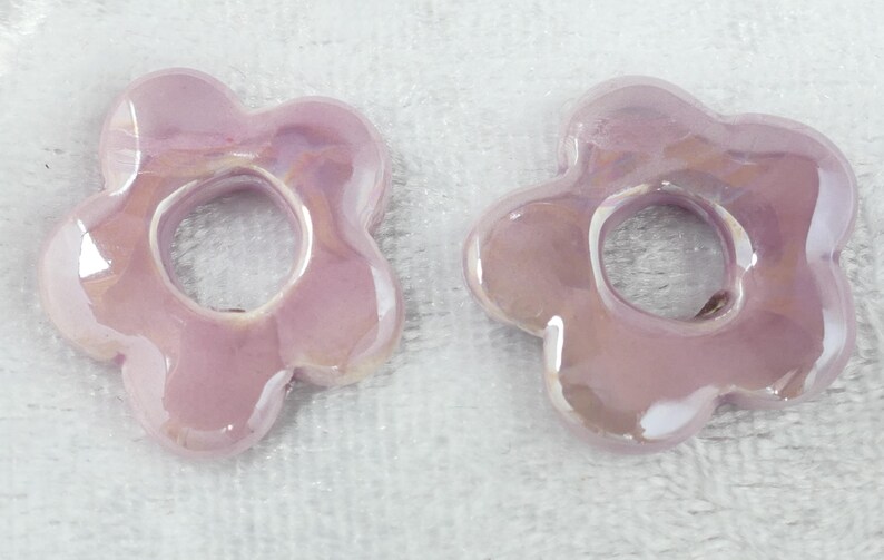 May include: Two iridescent, light purple flower-shaped ceramic beads. Each bead has a central hole and five rounded petals. The beads have a glossy finish and reflect light, creating a shimmering effect. They are set against a white background.