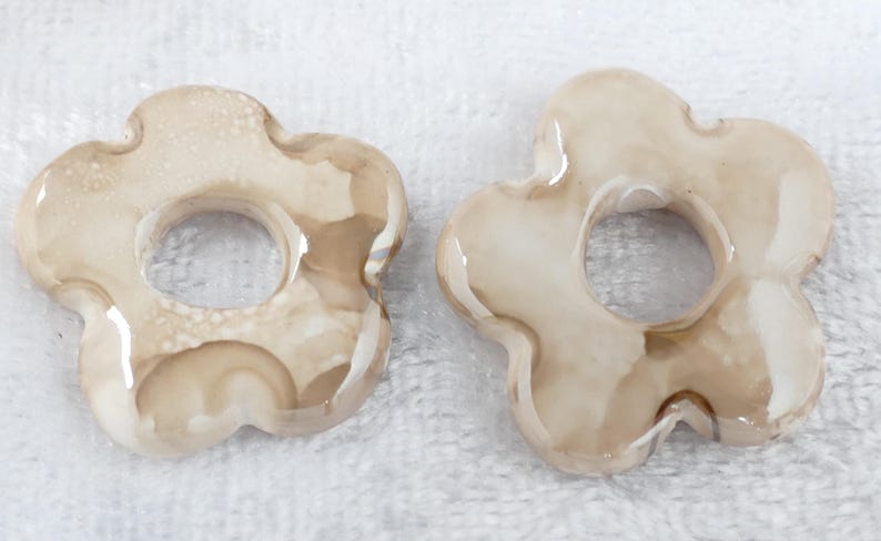 May include: Two flower-shaped decorative objects with a hole in the center. The objects are a light beige color with darker brown marbling. They have a glossy finish and are set against a white background.