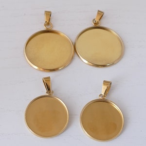 Pode incluir: Quatro bandejas de pendente de metal de cor dourada, de forma redonda e superfície lisa. As bandejas têm um laço na parte superior para prender a uma corrente ou cordão.