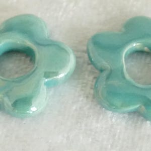 May include: Two turquoise ceramic flower-shaped beads with a glossy finish. Each bead has a central hole and petal-like edges. The beads are displayed on a white, textured surface, showcasing their color and design.