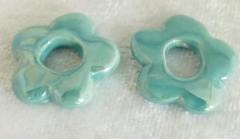 May include: Two turquoise ceramic flower-shaped beads with a glossy finish. Each bead has a central hole and petal-like edges. The beads are displayed on a white, textured surface, showcasing their color and design.