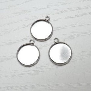Pode incluir: Três bandejas redondas para pingentes de cor prateada com um acabamento liso e brilhante. Cada bandeja tem um laço na parte superior para prender uma corrente ou um cordão.