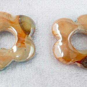 May include: Two flower-shaped beads with a hole in the center. The beads have a glossy finish with shades of orange, brown, and cream. The beads are likely made of glass or a similar material.