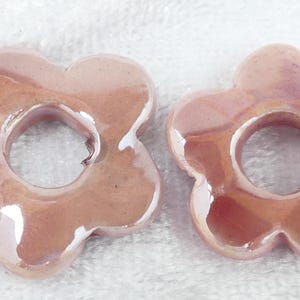 May include: Two flower-shaped ceramic beads in a soft pink hue with iridescent highlights. Each bead has a central hole and petal-like edges. The beads are likely for jewelry making or craft projects.
