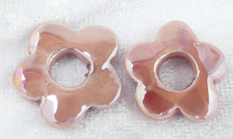 May include: Two flower-shaped ceramic beads in a soft pink hue with iridescent highlights. Each bead has a central hole and petal-like edges. The beads are likely for jewelry making or craft projects.