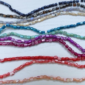 Può includere: Assortimento di perline quadrate colorate realizzate in conchiglia iridescente. Le perline sono infilate su una superficie bianca.