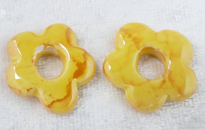 May include: Two yellow flower-shaped beads with a glossy finish. The beads have a hole in the center and a marbled effect with brown and orange hues. These beads are suitable for jewelry making.