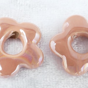 May include: Two flower-shaped beads in a soft peach color with an iridescent sheen. Each bead has a central hole and a glossy finish, suggesting they are made of ceramic or glass. The beads are likely for jewelry making.