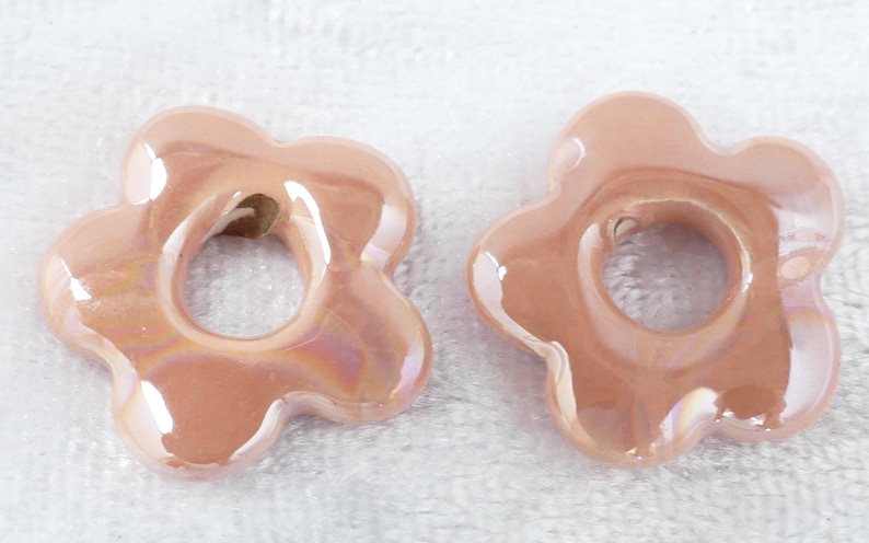 May include: Two flower-shaped beads in a soft peach color with an iridescent sheen. Each bead has a central hole and a glossy finish, suggesting they are made of ceramic or glass. The beads are likely for jewelry making.
