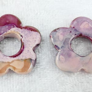 May include: Two flower-shaped ceramic beads with a hole in the center. The beads have a glossy finish and are primarily shades of pink, purple, and white. The floral design is a unique, handmade craft.