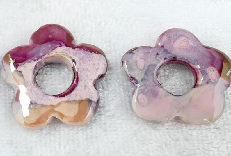 May include: Two flower-shaped ceramic beads with a hole in the center. The beads have a glossy finish and are primarily shades of pink, purple, and white. The floral design is a unique, handmade craft.