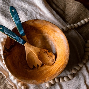 May include: A wooden salad bowl with two matching serving spoons. The spoons have teal and white epoxy resin handles. The bowl and spoons are made of light-coloured wood with a natural grain. The items are arranged on a neutral-coloured cloth.