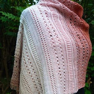 May include: A handmade crochet shawl in a gradient of colors, transitioning from light beige to a warm reddish-brown. The shawl features intricate openwork patterns and a fringed edge, displayed on a mannequin.