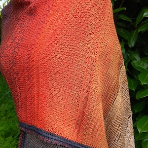 May include: A hand-knitted shawl with a gradient of orange, brown, and grey colours. The shawl has a crocheted border and a lacy pattern.