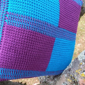 May include: A handmade crochet bag in a patchwork design. The bag features squares of bright turquoise and deep purple yarn. The bag is resting on a tree branch, showcasing the texture and color of the crochet work.