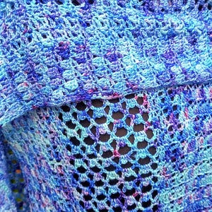 May include: A close-up of a handmade, open-weave shawl in shades of blue, purple, and teal. The crochet pattern creates a delicate, lacy texture. The shawl is made of soft yarn, perfect for layering. The color palette is vibrant and eye-catching.