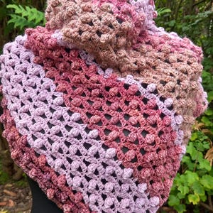 May include: A handmade crochet shawl in shades of pink, mauve, and brown. The shawl features a textured, openwork design with a triangular shape. The item is displayed on a black mannequin.