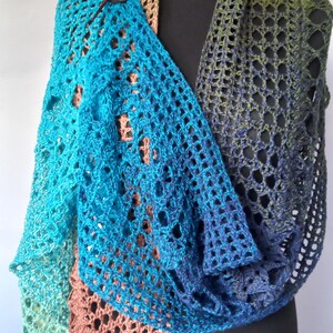 May include: A crochet shawl featuring a gradient of colors including light green, turquoise, blue, and copper. The openwork fabric is secured at the shoulder with a dark brown wooden toggle pin. The shawl is draped over a black display form.