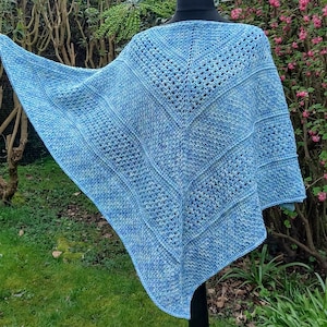 May include: A light blue crocheted shawl displayed on a black mannequin. The shawl features a geometric pattern with varying textures and shades of blue. The open weave design adds visual interest. The background includes green foliage and pink flowers.