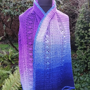 May include: A crocheted shawl with a gradient of colors from white to purple. The shawl is draped over a mannequin and has a delicate lace pattern.