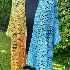 Op de afbeelding: Een gehaakte omslagdoek met een kleurgeblokt ontwerp, waarbij de ene kant mosterdgeel is en de andere kant lichtblauw. Het kledingstuk heeft een ingewikkeld opengewerkt kantpatroon en een verfijnde geschulpte rand langs de onderkant.