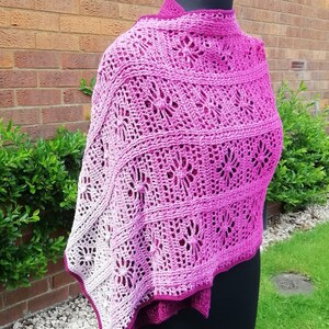 May include: A handmade crochet shawl in shades of pink and white, draped over a mannequin. The shawl features a floral openwork design and a scalloped edge. The color gradient transitions from a deep magenta to a soft white.