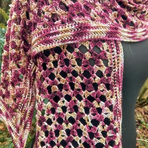 May include: A crocheted shawl in a burgundy, beige, and white color scheme. The shawl has a lacy pattern and a fringed edge.