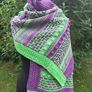 May include: A handmade, knitted shawl in shades of purple and green. The shawl features a textured pattern with a green border and accents. The design includes diagonal stripes and textured bobbles, creating a visually appealing accessory.