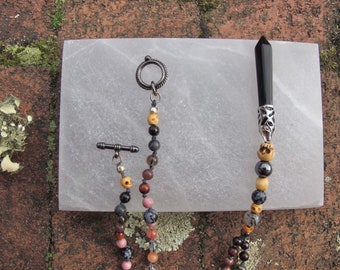 May include: A multi-strand beaded necklace with a black obsidian point pendant. The necklace features a variety of beads, including black, brown, gray, and white beads. The necklace is displayed on a white surface.