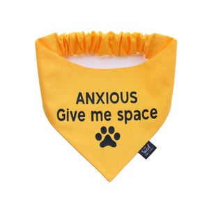 May include: Yellow dog bandana with the text "ANXIOUS Give me space" in black, along with a black paw print. The bandana has an elastic neckband. A small black tag is attached to the side.