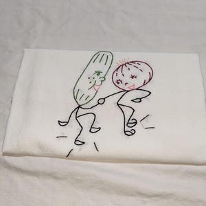 May include: A white kitchen towel featuring an embroidered design of a green cucumber and a red onion with stick-figure arms and legs. The two vegetables are depicted holding hands and dancing, with black stitched lines indicating movement.