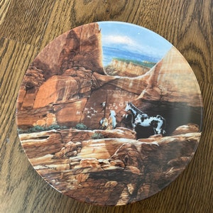 May include: A round ceramic plate with a scenic image of a canyon with a blue sky and a herd of horses. The plate has a brown, orange, and white color scheme.