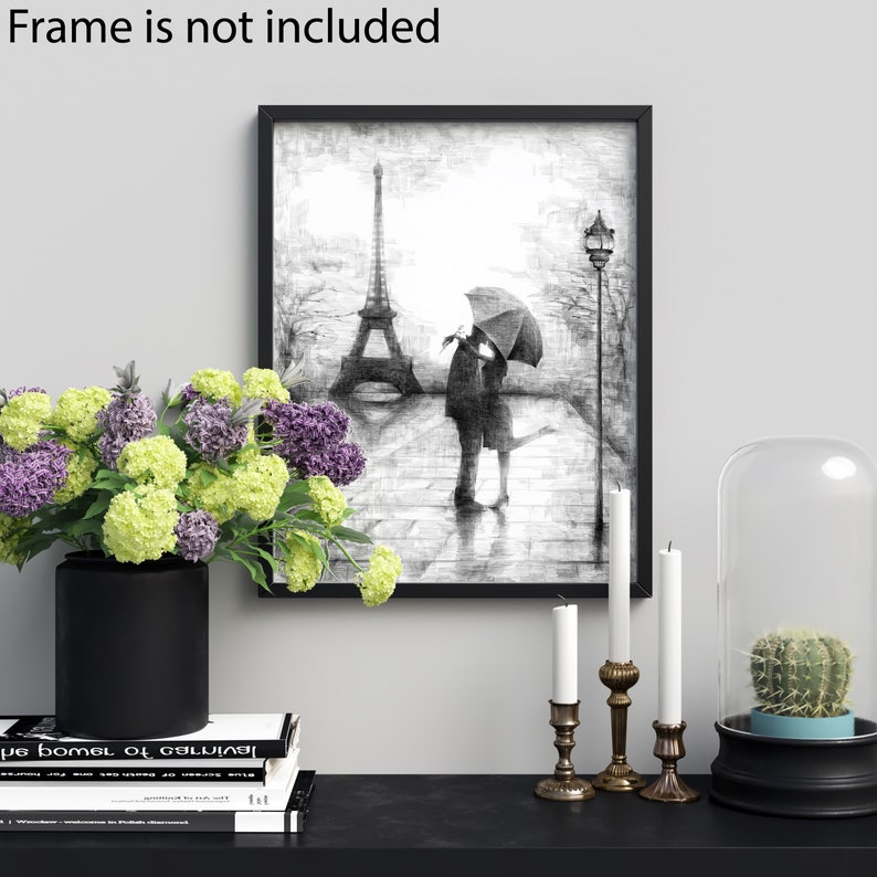 Pode incluir: Desenho a l&aacute;pis preto e branco de um casal se beijando sob um guarda-chuva em frente &agrave; Torre Eiffel. O desenho est&aacute; emoldurado e exposto em uma prateleira preta com um vaso de flores, velas e uma redoma de vidro com um cacto.