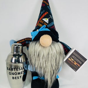Pu&ograve; includere: Un gnomo di peluche che indossa un cappello nero e blu con una stampa di cocktail. Il gnomo tiene uno shaker per cocktail argentato che dice "Bartesian Gnomes Best". Il gnomo tiene anche una piccola scatola bianca che dice "Bartesian The Classic Collection".
