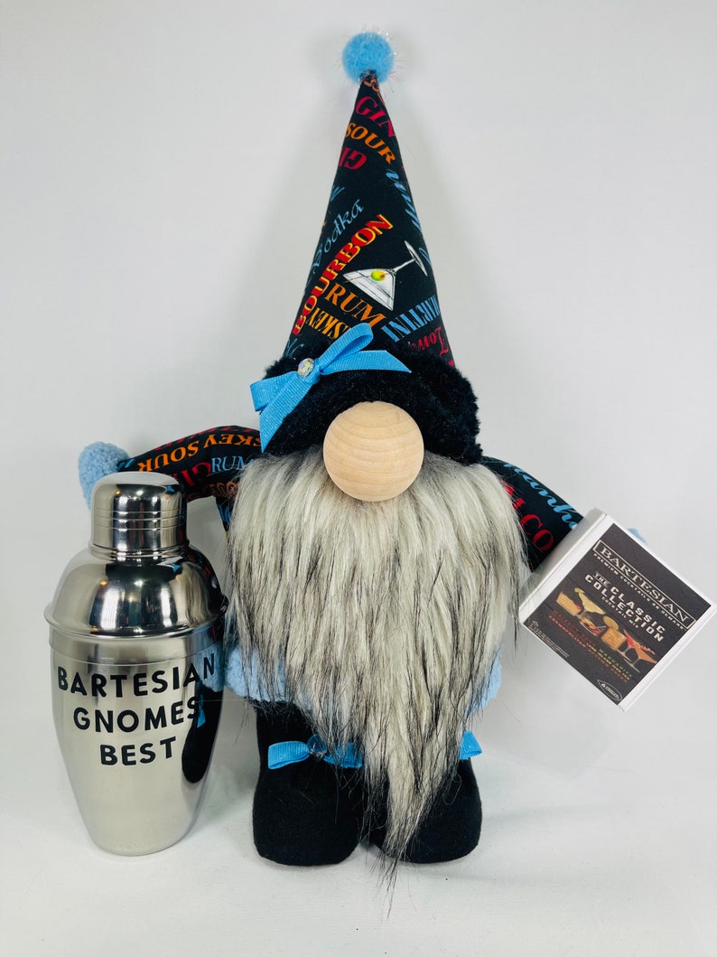 Pu&ograve; includere: Un gnomo di peluche che indossa un cappello nero e blu con una stampa di cocktail. Il gnomo tiene uno shaker per cocktail argentato che dice "Bartesian Gnomes Best". Il gnomo tiene anche una piccola scatola bianca che dice "Bartesian The Classic Collection".