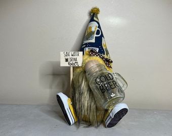 Peut inclure: Un gnome jaune et noir portant un chapeau bleu et blanc sur le thème de la bière. Le gnome tient un bocal en verre avec les mots "Cold Beer" écrits dessus. Le gnome tient également une pancarte qui dit "Save Water Drink Beer".